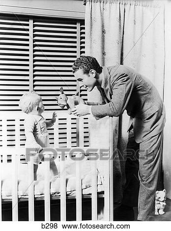 1930S 1940S 11 Month Old Baby Standing In Crib In Front Of Window With Venetian Blinds Father Handing Baby A Toy Dog Stuffed Animal Nursery View Large Photo Image Stock Photo - 1930S 1940S 11 Month Old Baby Standing In Crib In Front Of Window With Venetian Blinds Father Handing Baby A Toy Dog Stuffed Animal Nursery. Fotosearch
