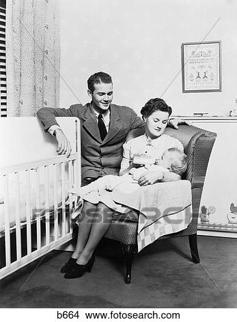 1930S 1940S Mother Sitting In Chair In Nursery Baby 11 Months On Lap Drinking From Bottle Father Looking On Leaning On Crib View Large Photo Image Picture - 1930S 1940S Mother Sitting In Chair In Nursery Baby 11 Months On Lap Drinking From Bottle Father Looking On Leaning On Crib. Fotosearch