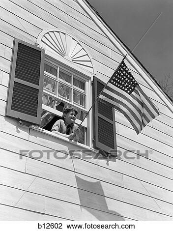 Stock Image - 1930S 1940S Smiling Boy Leaning Out Of Upstairs Window Holding American Flag. Fotosearch