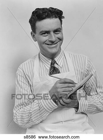 Stock Photograph - 1930S 1940S Smiling Curly Hair Man Wearing Apron Taking Order With Pencil And Note Pad Looking At Camera. Fotosearch