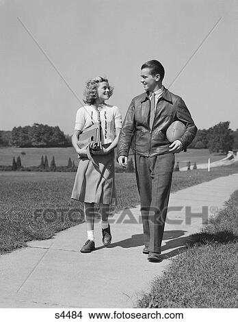 Picture - 1930S 1940S Teen Couple Walking Down Sidewalk Girl With School Books Boy Holding Football . Fotosearch
