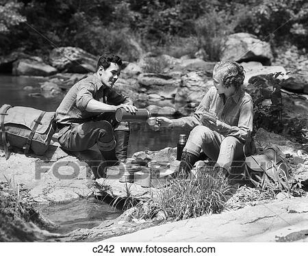 Stock Image - 1930S Couple Having Picnic Sitting On Rocks By Stream Man Pouring Drink For Woman From Thermos. Fotosearch