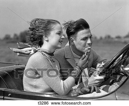 Stock Photograph - 1930S Man Driving Convertible Automobile While Girlfriend Lights His Cigarette. Fotosearch