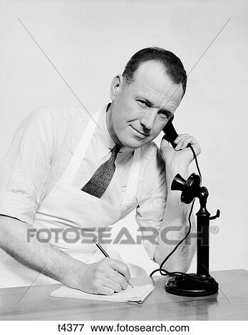 1930S Portrait Man Clerk Wearing Apron Talking On Old Black Upright Candlestick Telephone Holding Ear Piece To Ear Writing On Pad With Pencil Looking At Camera View Large Photo Image Stock Photo - 1930S Portrait Man Clerk Wearing Apron Talking On Old Black Upright Candlestick Telephone Holding Ear Piece To Ear Writing On Pad With Pencil Looking At Camera. Fotosearch