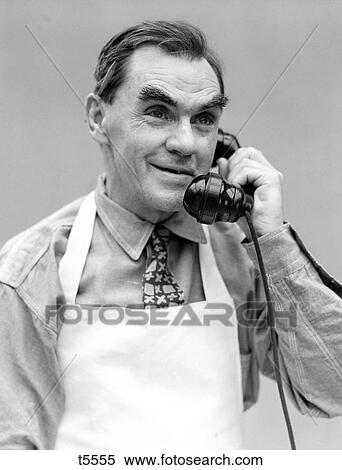 Stock Photography - 1930S Portrait Older Man Wearing Apron Talking On Telephone Pencil Behind Ear Smiling Bushy Eye Brows. Fotosearch