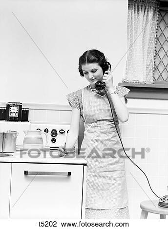 Stock Image - 1930S Woman Housewife Standing Kitchen By Stove Talking On Telephone Writing On Pad Wearing Apron . Fotosearch
