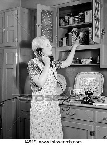 1930S Woman Housewife Talking On Black Rotary Cradle Telephone Looking Into Kitchen Cupboard View Large Photo Image Stock Photo - 1930S Woman Housewife Talking On Black Rotary Cradle Telephone Looking Into Kitchen Cupboard . Fotosearch