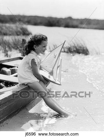 1930S Young Girl At Seashore Holding Sailboat Toy Sitting On Edge Of Rowboat Feet In Water View Large Photo Image Stock Photograph - 1930S Young Girl At Seashore Holding Sailboat Toy Sitting On Edge Of Rowboat Feet In Water. Fotosearch