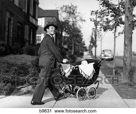 Stock Image - 1940S 1950S Smiling Father In Suit & Hat Pushing Baby In Carriage Down Sidewalk. Fotosearch