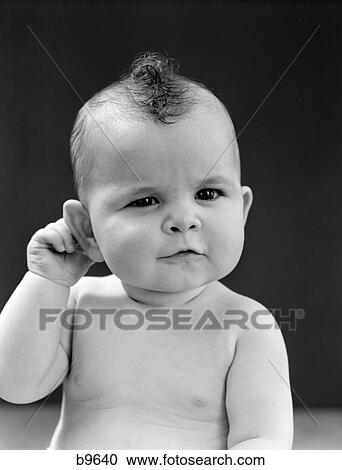 Stock Image - 1940S Baby With Slight Squinting Eyes And Puckered Mouth Holding Right Hand Behind Right Ear Studio. Fotosearch