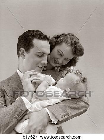 Stock Photo - 1940S Father Cradling Baby Daughter Feeding Her Bottle With Mother Looking Over His Shoulder. Fotosearch
