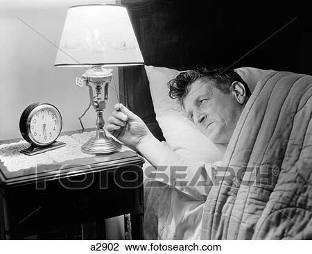 Stock Image - 1940S Tired Man In Bed Turning On Light Next To Alarm Clock Showing Time As 6. 30 Am. Fotosearch