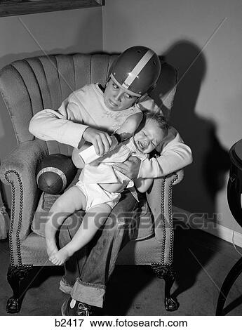 1950S 1960S Boy Sitting By Football In Easy Chair Wearing Helmet Trying To Hold And Feed Bottle To Crying Baby View Large Photo Image Stock Photo - 1950S 1960S Boy Sitting By Football In Easy Chair Wearing Helmet Trying To Hold And Feed Bottle To Crying Baby. Fotosearch