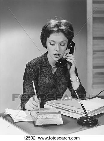 Stock Image - 1950S 1960S Brunette Business Woman Sitting Desk Talking Telephone Writing Note On Calendar. Fotosearch