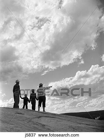 1950S 1960S Family Of Four Silhouetted On Top Rocky Hill Pointing At Clouds In Sky View Large Photo Image Stock Photo - 1950S 1960S Family Of Four Silhouetted On Top Rocky Hill Pointing At Clouds In Sky. Fotosearch