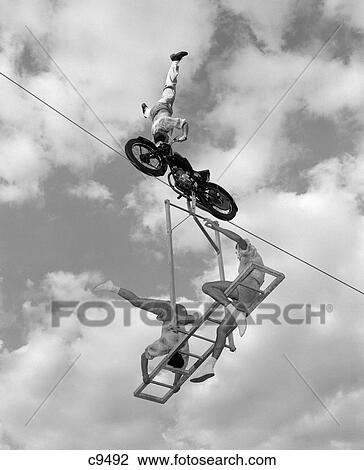 Stock Image - 1950S 1960S High-Wire Act With Man Standing On His Head On Motorcycle With Man And Woman On Balanced Ladder Suspended Below. Fotosearch