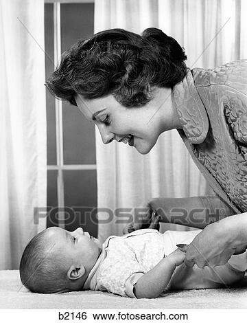 Stock Photograph - 1950S 1960S Mother Smiling At Baby Lying On Table Indoor. Fotosearch