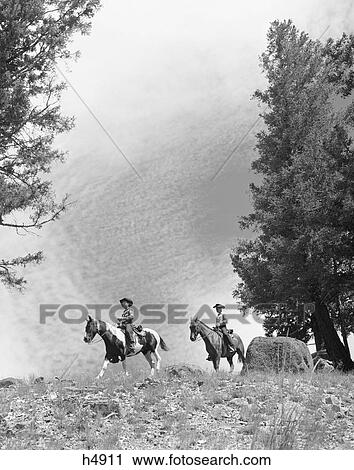 Stock Image - 1950S 1960S Two Men On Horseback Riding Across Field Wearing Cowboy Hats. Fotosearch