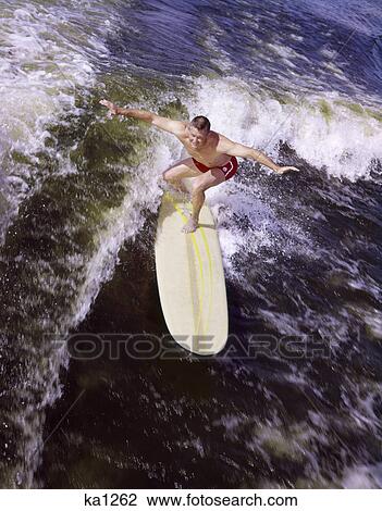 Stock Image - 1950S 1960S Young Man Red Swim Trunks Yellow Surfboard Riding A Wave Surfing . Fotosearch
