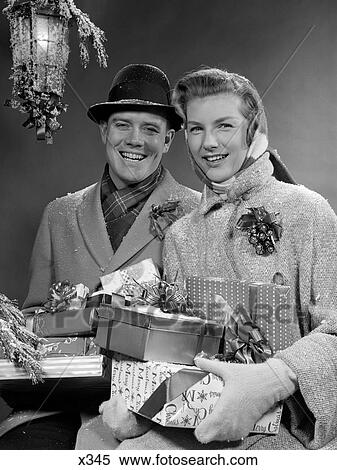 Stock Photography - 1950S Couple Husband And Wife Carrying Armload Of Christmas Packages Smiling Standing In Snow Under Porch Light Outdoor. Fotosearch