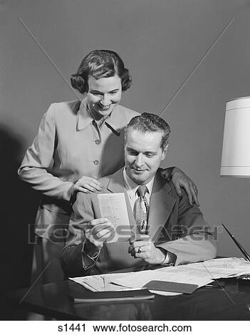 Stock Image - 1950S Couple Man Seated At Desk Holding Paper Woman Standing Looking Over His Shoulder Smiling Happy. Fotosearch