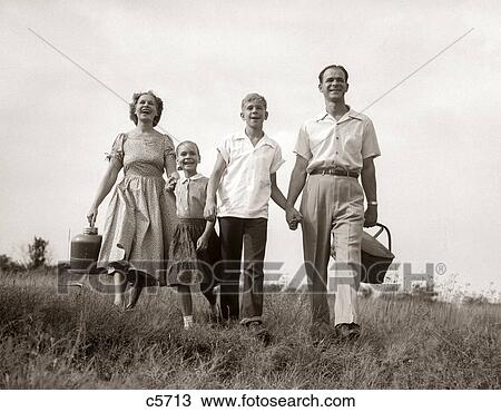 Stock Image - 1950S Family Walking In Grassy Meadow Carrying Summer Picnic Basket And Thermos. Fotosearch