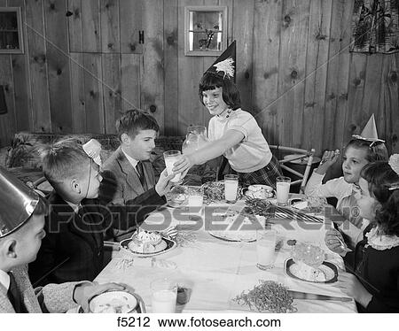 Stock Image - 1950S Girl Pouring Glasses Milk For Group Children At Birthday Party Wearing Party Hats Eating Cake. Fotosearch
