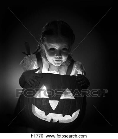 Stock Image - 1950S Little Girl Standing Over Carved Pumpkin Face Lit By Candle. Fotosearch