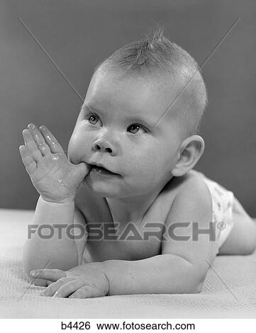Stock Photograph - 1950S Portrait Baby Lying On Stomach With Thumb In Mouth. Fotosearch