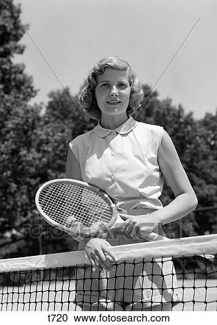 Stock Image - 1950S Portrait Woman Holding Tennis Racquet Standing Behind Net Outdoor Looking At Camera. Fotosearch