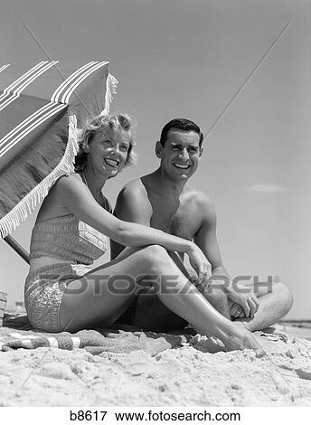 Stock Photo - 1950S Smiling Couple Sitting On Beach With Umbrella. Fotosearch