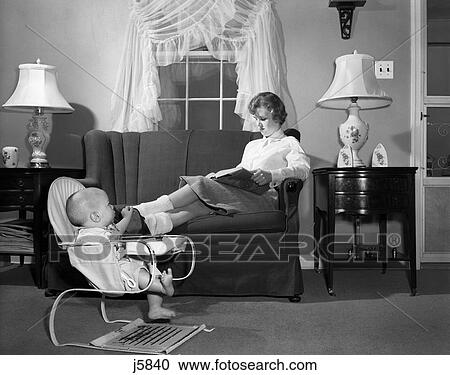 Stock Image - 1950S Teen Babysitter Seated On Sofa Reading Schoolbook While Baby In Bouncy Sling Chair Grabs Her Propped Up Foot. Fotosearch
