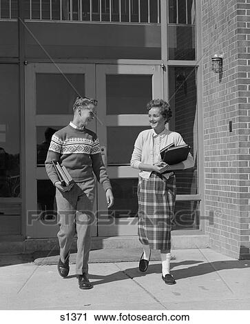 Stock Image - 1950S Two Teenage Students Boy And Girl Walking Away From School Doors Carrying Books . Fotosearch