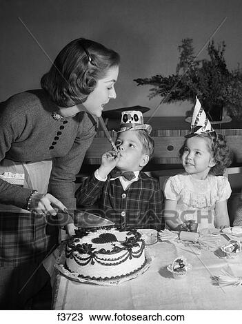 Stock Image - 1950S Woman Mother Cutting Birthday Cake For Two Children Sitting At Table Wearing Party Hats. Fotosearch