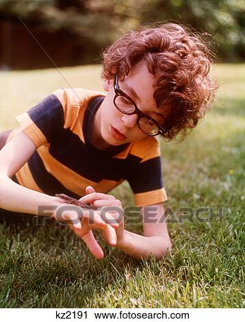 1960S 1970S Geeky Boy Wearing Striped T-Shirt Big Eye Glasses Lying In Backyard Grass Playing With Pet Reptile Lizard View Large Photo Image Stock Image - 1960S 1970S Geeky Boy Wearing Striped T-Shirt Big Eye Glasses Lying In Backyard Grass Playing With Pet Reptile Lizard. Fotosearch