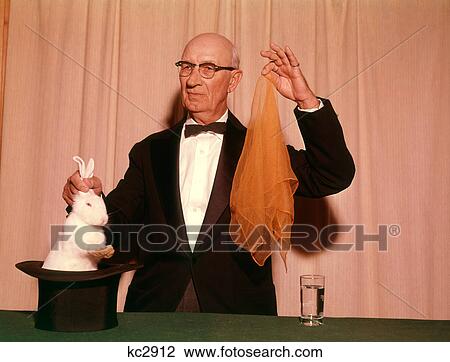Stock Image - 1960S 1970S Magician Pulling A White Rabbit Out Of Hat And Holding An Orange Scarf. Fotosearch