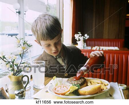 Stock Photograph - 1960S Boy Eating At Booth In Restaurant Pouring Ketchup On Hamburger. Fotosearch