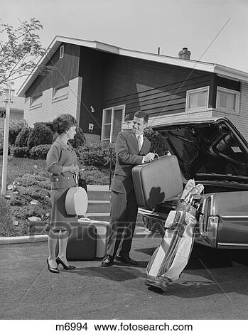 Picture - 1960S Couple Man Woman Packing Luggage Into Car Trunk By Suburban House. Fotosearch
