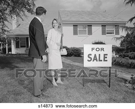 Stock Image - 1960S Couple Man Woman Standing In Front Of House With For Sale Sign In Yard. Fotosearch