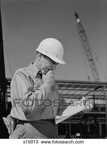Stock Image - 1960S Male Construction Site Supervisor Wearing Hard Hat Reading Papers. Fotosearch
