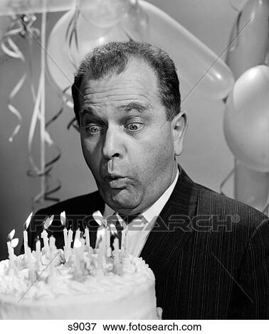 Stock Photo - 1960S Man Blowing Out Candles On Birthday Cake With Bulging Eyes & Balloons In Background. Fotosearch