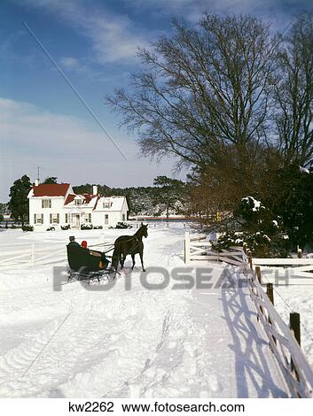 Stock Image - 1960S Man Woman Couple In One Horse Drawn Open Sleigh Approaching Snowy White Farm House. Fotosearch
