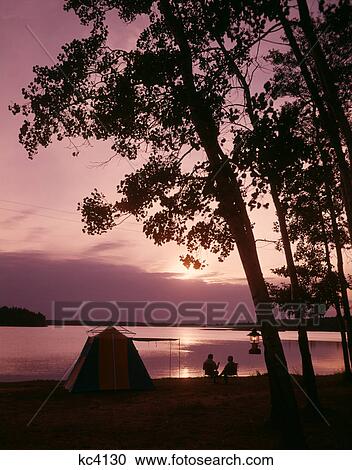 Stock Image - 1960S Silhouette Of Couple Camping By Lake At Sunset. Fotosearch