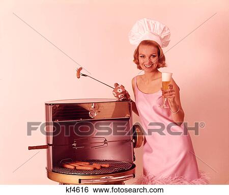 Stock Photograph - 1960S Smiling Blond Woman Wearing Pink Party Dress And Chef Hat Grilling Hot Dogs Drinking Beer. Fotosearch