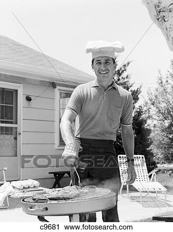 Stock Image - 1960S Smiling Man Outdoors In Backyard Patio Wearing Chef Hat Cooking Steaks Hot Dogs On Grill. Fotosearch