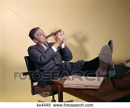 1960S Successful Businessman With Look Of Satisfaction Sitting With Shoes Propped On Desk Lighting-Up A Huge Cigar View Large Photo Image Stock Photography - 1960S Successful Businessman With Look Of Satisfaction Sitting With Shoes Propped On Desk Lighting-Up A Huge Cigar. Fotosearch