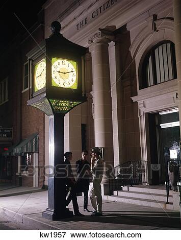 Stock Photo - 1960S Three Teenage Boys Loitering Under Clock Outside Bank Building Smoking Cigarettes At Night. Fotosearch