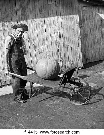 Stock Photography - 1970S 1980S Elderly Farmer In Overalls & Straw Hat Looking At Camera Pushing Old Wheelbarrow With Huge Pumpkin On It. Fotosearch