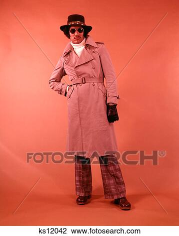 Stock Image - 1970S African American Man In Tan Trench Coat Aviator Sunglasses Hat Gloves Turtleneck Plaid Bellbottom Plaid Pants Looking At Camera. Fotosearch