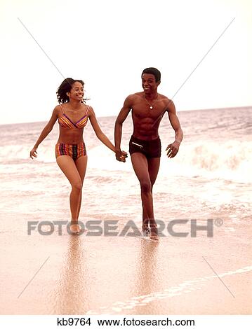 Picture - 1970S African American Young Couple Wearing Bathing Suits Running In Ocean Surf. Fotosearch
